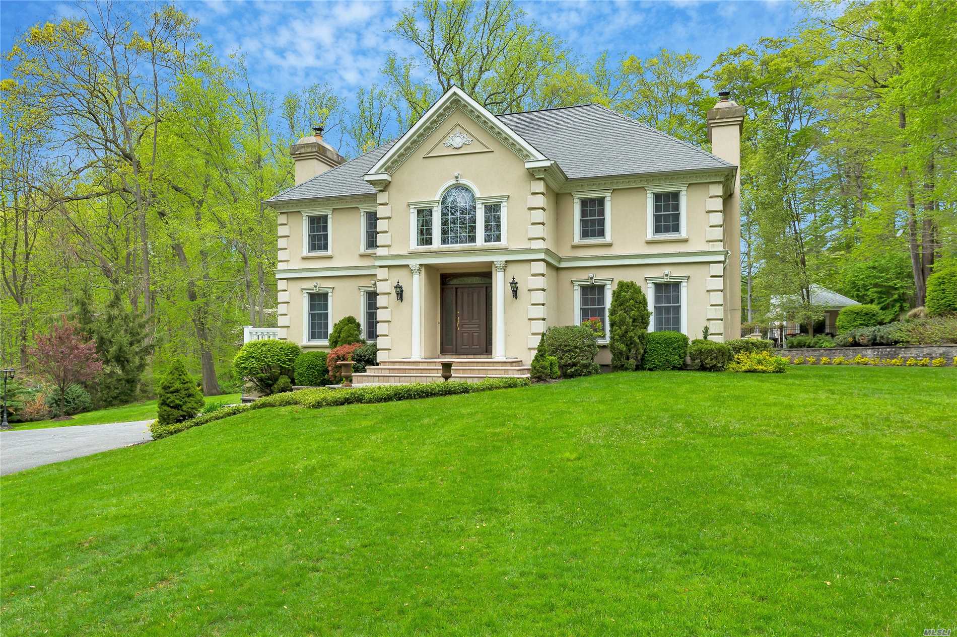 Standing regally on a gentle hillside, this timeless stucco French Colonial, with coins, hip roof, domed chimneys, elegant moldings, and coat-of-arms, welcomes guests up a long, winding drive partially shaded by stately, towering oaks. Off a quiet cul-de-sac in Lattingtown, this magnificent two-acre mini-estate is carved out of pristine woodland and features sweeping emerald lawns, stone-tiered gardens, and an entertainer’s-dream outdoor entertainment complex. An enthralling country-club ambiance carries on into the home’s bright and spacious, four-bedroom, three-bath interior offering a casual elegance with easy flow from space to space, natural-tone hardwood floors, elegant millwork, large windows, French doors, a fabulous finished basement, and a huge unfinished attic with expansion possibilities. On the shores of Long Island Sound, Lattingtown is the northernmost part of a larger area, fanning out from the village of Locust Valley, that is referred to by many as “Locust Valley,” just as all seaside communities on Long Island’s lower eastern peninsula are called “the Hamptons.” Located less than 30 miles from Manhattan, it has in recent years become a closer and more appealing destination for a younger generation of successful New Yorkers to live.  It is known for its grand estates, scenic vistas, country and golf clubs, and its proximity to yacht clubs, fine dining, quaint shopping districts, private and public schools, and the Long Island Railroad. 
Amidst serene gardens and rolling lawns with a backdrop of soaring trees, the rear property’s resort-like retreat begins with an expansive rear granite patio, ideal for al fresco dining, that leads via a bluestone path through gated estate-fencing to an enchanting inground, Gunite, salt-water pool that has been recently renovated like new. Hidden pool equipment includes a brand-new filter system with a super-quiet, four-speed pump, salt cell, and existing heater. Upgraded electrical equipment comprises six Intellibrite LED pool lights with multi-color functions and software for manual or phone/tablet control of lights, pump, heater, and waterfalls. An expansive surround and patio are enhanced by a new blue-tile raised wall containing a triple waterfall. A brand-new pool house, designed, architecturally drawn, and built from the ground up, boasts central-air conditioning and heat for year-round enjoyment. A large great room contains a granite-topped kitchenette with center island, high-end refrigerator, freezer drawers, ice machine cabinet, and dishwasher. There is a full bath with a glass enclosed indoor shower plus an outdoor shower with its own brand-new septic system. A Nana window system opens the kitchen to a six-foot bar, with ample seating, in one of two raised verandas overlooking the pool.
The interior begins with a two-story central foyer, with elegant staircase, extending through to kitchen and French doors accessing the rear deck. The living room is flooded with light through two six-over-nine front windows and French doors to a side balcony flanking a massive marble fireplace. The formal dining room opens through wide French doors to a versatile sitting room, with direct access to the kitchen, ideal for additional seating or buffet for large dinner parties. The sky-lit, vaulted-ceiling kitchen enjoys custom cabinetry with white marble countertop, high-end appliances, a farm sink overlooking the patio, a pantry, and a delightful breakfast area offering gorgeous vistas through a bay of six large windows. A hall off the foyer leads to a lovely marble full bath and a spacious study/family room with fireplace and French doors to the rear patio. This area could easily convert to a first-floor master suite if desired. 
The second-floor master suite offers a large bedroom with ample closets and room for more, a gorgeous marble master bath with whirlpool tub and glass-enclosed shower, and access to a private balcony overlooking the foyer with Palladian window and two closets. Two additional bedrooms share an attractive hall bath. The fabulous finished basement is ideal for entertaining and has access to the driveway. It includes a great room with fireplace, game room, gym, bonus/sound room, bedroom, full bath, and access to the attached two-car garage. Offering the quintessence of “Gold Coast” luxury living, this exceptional pristine home is ready for you to make it your own Shangri-la.